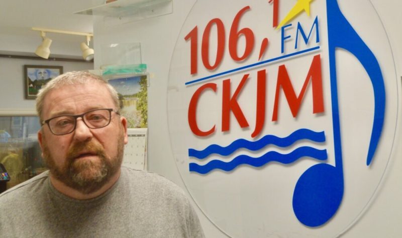 Un homme avec des lunettes et une barbe portant un chandail brun en avant du logo de Radio CKJM.