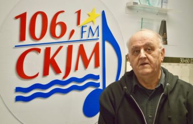 Un homme avec chemise et gilet noirs debout en avant du logo de Radio CKJM.