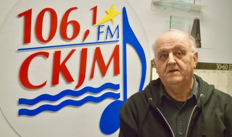 Un homme avec chemise et gilet noirs debout en avant du logo de Radio CKJM.