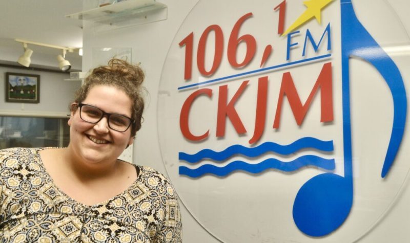 Une dame avec des lunettes et en chandail vert et jsune en avant du logo de Radio CKJM.