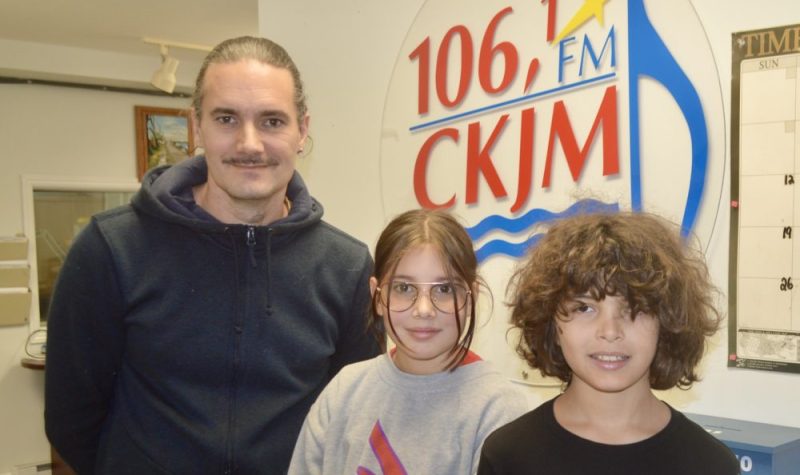 Un homme et deux jeunes en avant du logo de Radio CKJM.