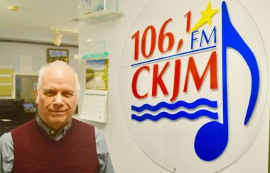 Un homme portant une chemise grise et une vest violette en avant du logo de Radio CKJM.