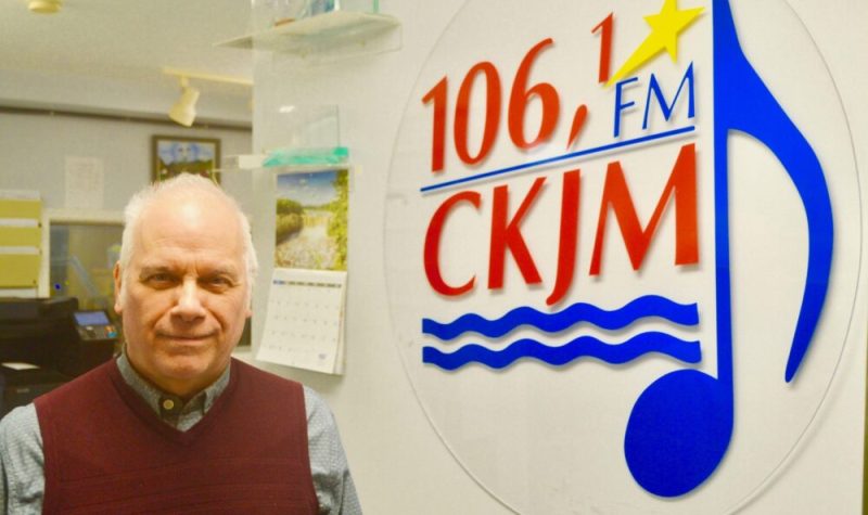 Un homme portant une chemise grise et une vest violette en avant du logo de Radio CKJM.