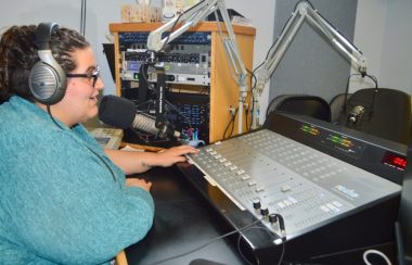 Dame avec chandail bleu et des écouteurs parlant dans un micro en avant d'une console de radio.