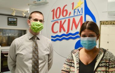 Homme avec chemise et cravate brune portant un masque vert et femme avec chandail multicolore en avant du logo de Radio CKJM.