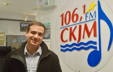 Homme avec chemise rose et gilet brun en avant du logo de Radio CKJM.