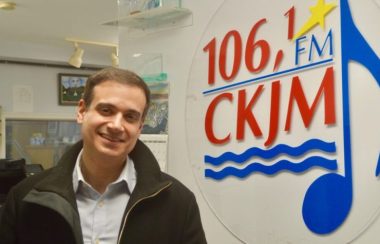 Un homme portant une chemise bleue et un gilet noir en avant du logo de Radio CKJM.
