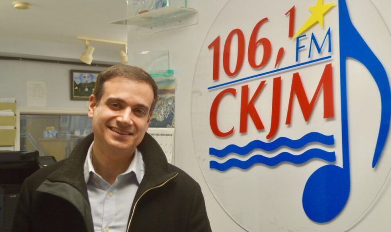 Un homme portant une chemise bleue et un gilet noir en avant du logo de Radio CKJM.