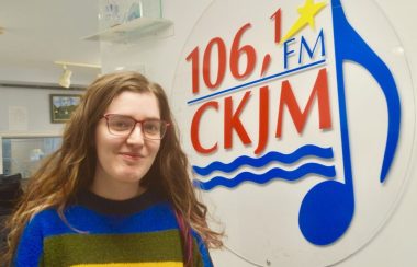 Jeune fille aux cheveux longs portant un chandail bleu, vert et jaune en avant du logo de Radio CKJM.