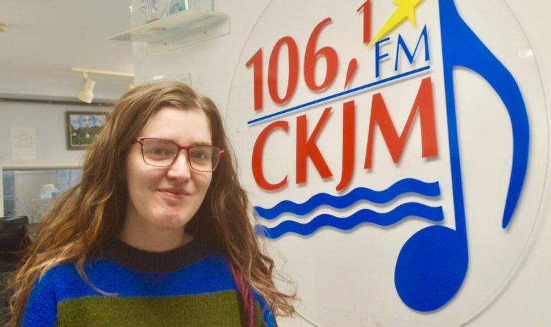 Jeune fille aux cheveux longs portant un chandail bleu, vert et jaune en avant du logo de Radio CKJM.