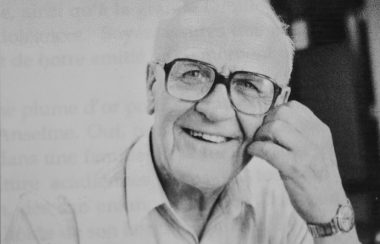 Homme âgé avec lunettes et chemise blanche souriant à la caméra.