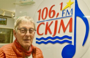 Homme avec lunettes et gilet orange en avant d'un logo d'un poste de radio.