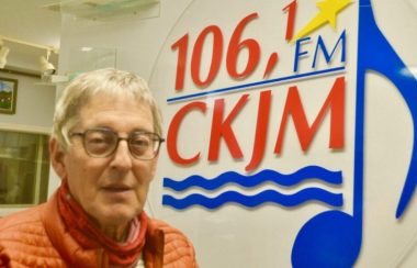 Un homme portant un manteau orange en avant du logo de Radio CKJM.