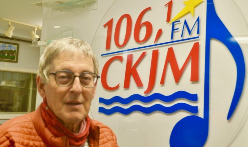 Homme avec lunettes et gilet orange en avant d'un logo d'un poste de radio.