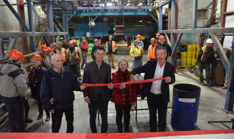 Four officials stand with their hands on a ribbon, with the one on the right holding a pair of scissors while workers in hard hats watch in the background.