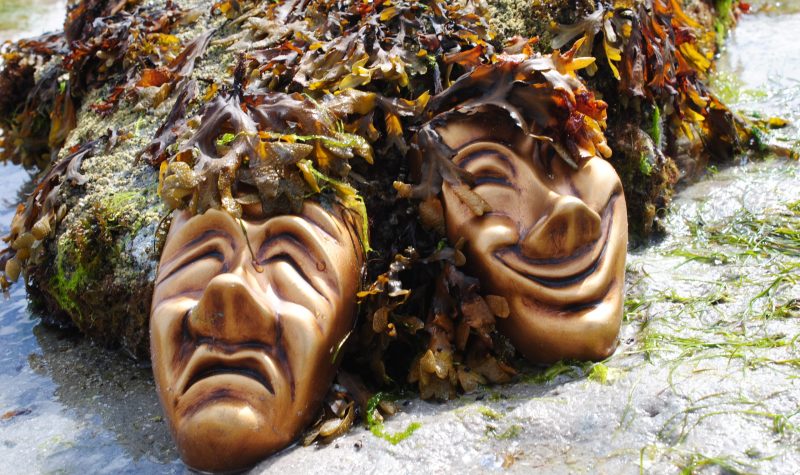 Two dramatic masks rest on a pile of seaweed on the beach.