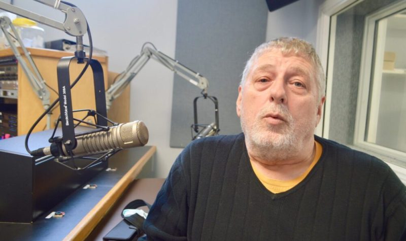 Homme avec chandail noir assis à côté d,un micro dans un studio de radio.