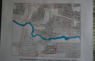 A black and white map shows a small village with a small blue river running through it and a red outline in a smaller portion of the village. The red outline marks the boundary of the actual Police Village of Russell.