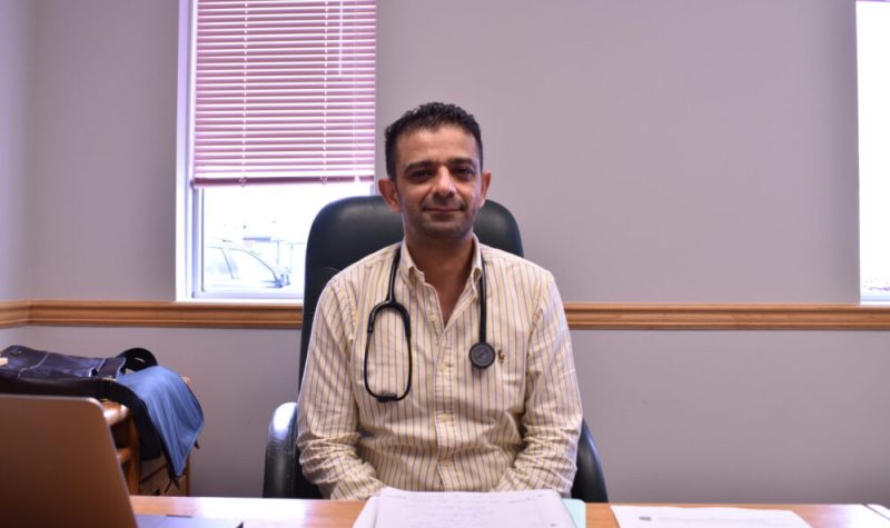 Dr Hussein sourit, assis à son bureau sur lequel sont étalés des papiers et un ordinateur.