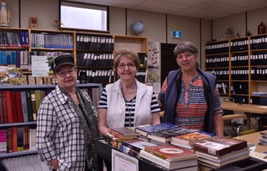 Trois personnes, Yvonne Desaulniers Cruikshank, Lucille Bussière et Paulette Desaulniers Briand se tiennent devant une table couverte de livres et derrière des étagères remplies de livres.