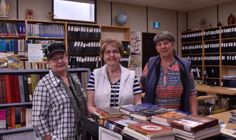 Trois personnes, Yvonne Desaulniers Cruikshank, Lucille Bussière et Paulette Desaulniers Briand se tiennent devant une table couverte de livres et derrière des étagères remplies de livres.