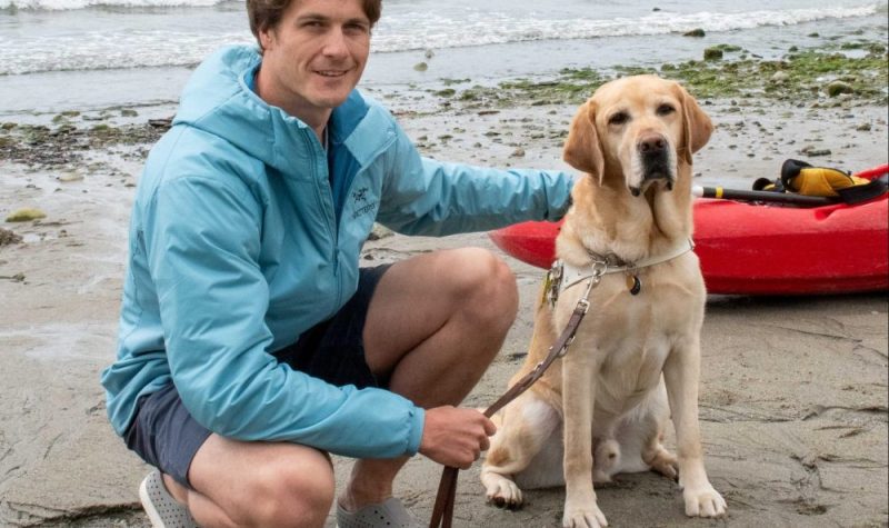 A man wearing a blue athletic jacket and shorts kneeling beside a yellow lab on a beach with a red kayack in the background.