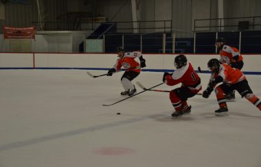 A hockey forward in a red jersey and wearing a cage tries to get past three defenders wearing orange and white jerseys.