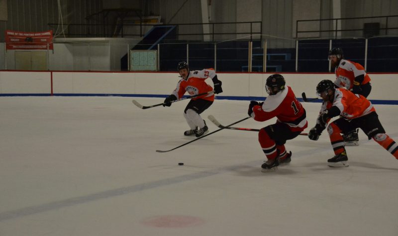 A hockey forward in a red jersey and wearing a cage tries to get past three defenders wearing orange and white jerseys.