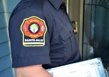 Un pompier de Sainte-Julie vêtu d'une chemise bleue foncée. Sur son épaule, l'armoirie du service de sécurité incendie de Sainte-Julie. Le pompier transporte une pile de papier.