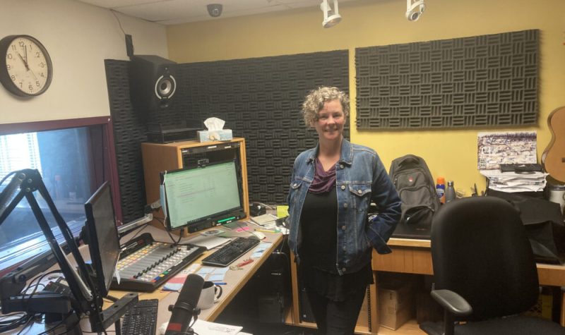 Une photo de Danielle Sturk dans les bureaux d'enregistrement de la radio Envol 91 FM. C'est une femme d'âge moyen aux cheveux argentés frisés et coupés à la garçonne. Elle port une veste en jean bleu sur un haut violet avec un jean bleu foncé et se tien au milieu du studio le sourire aux lèvres.