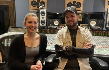 A woman and man stand side by side in a recording studio. There is a recording booth and equipment behind them.