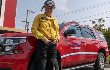 A man in firefighting gear stands in front of a red Nanaimo Fire Rescue SUV.