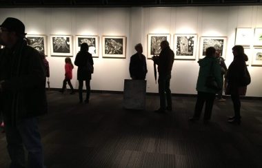 Plusieurs personnes assistent à une exposition. Six oeuvres sont exposées sur un mur blanc