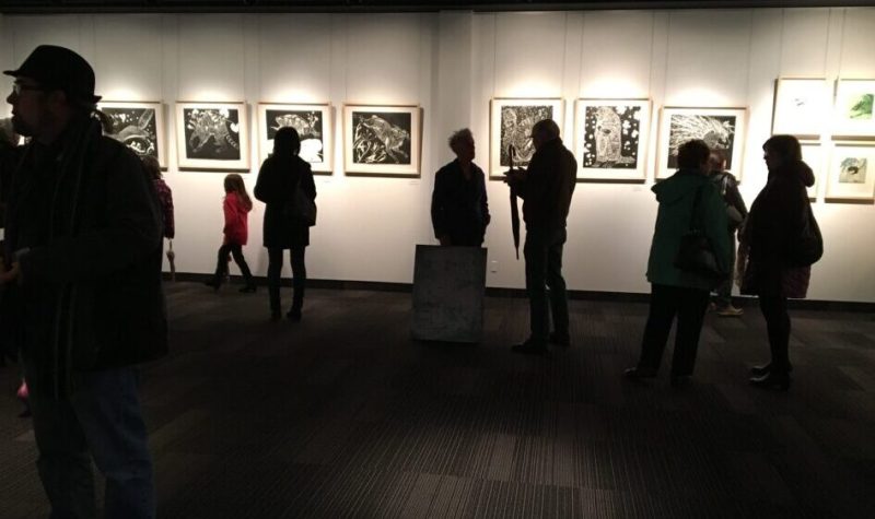 Plusieurs personnes assistent à une exposition. Six oeuvres sont exposées sur un mur blanc