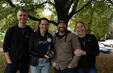 Four people stand shoulder to shoulder. A woman holds a BC Beer Award Trophy. They smile in celebration. A large deciduous tree serves as a backdrop behind them.