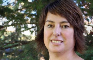 A headshot of a woman outside with green trees behind her on a sunny day.