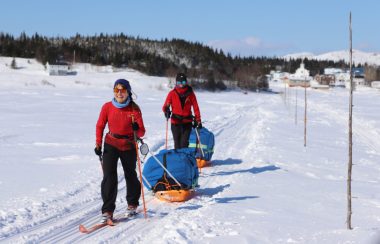 Une derrière l'autre, deux jeunes femmes en ski de fond tirent chacune un imposant traineau. Au loin, un petit village.