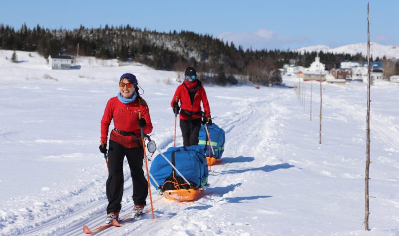 Une derrière l'autre, deux jeunes femmes en ski de fond tirent chacune un imposant traineau. Au loin, un petit village.