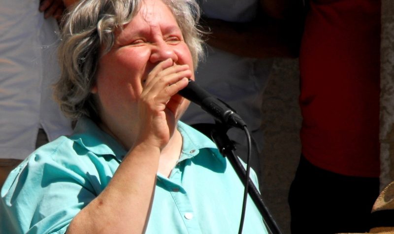 Denise Allard porte une chemise pastel et chante dans un microphone lors d'un événement organisé à l'extérieur.