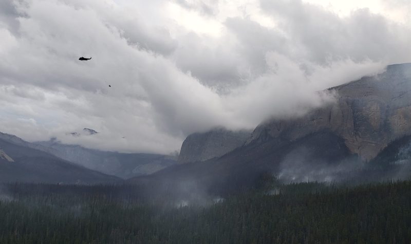 A helicopter angle of a forest fire happening on a ridge, while another helicopter fights the blaze. Weather is cloudy with lots of smoke in the air.