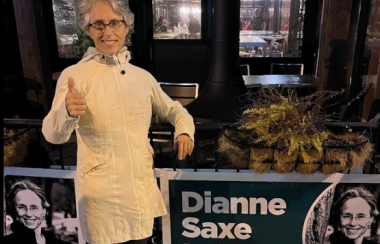 A person in a white jacket is giving a thumbs in front of signs and windows.