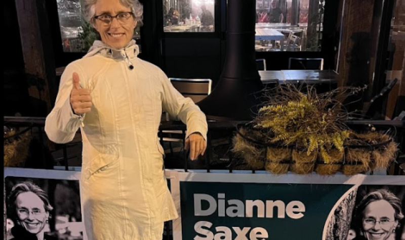 A person in a white jacket is giving a thumbs in front of signs and windows.