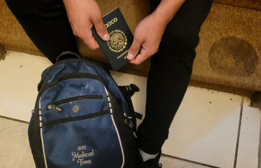 Un pasaporte de México sostenido por un par de manos, tambi`pen se ve una mochila junto a unos pies