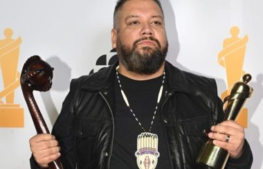 DJ Shub poses for pictures holding a juno award in one hard and a traditional war club artifact in the other. Standing in front of a white backdrop.