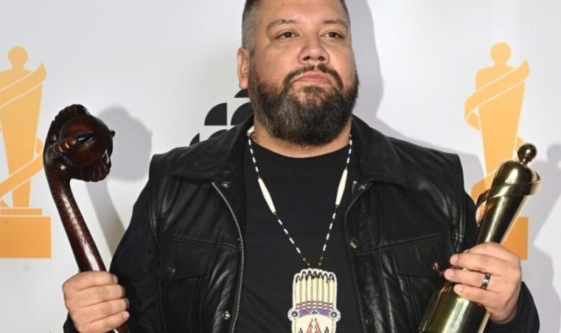 DJ Shub poses for pictures holding a juno award in one hard and a traditional war club artifact in the other. Standing in front of a white backdrop.
