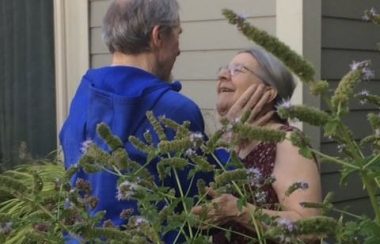 A man wearing a blue jacket holds the face of a woman outside a care home. There is a tree in front.