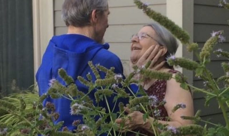 A man wearing a blue jacket holds the face of a woman outside a care home. There is a tree in front.