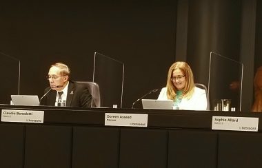 Le conseiller de Brossard Claudio Benedetti et la mairesse Doreen Assaad sont assis côte à côte à la table du conseil municipal. Monsieur Benedetti est vêtu d'un complet noir, d'une chemise blanche et d'une cravate rouge. Madame Assaad a un chandail turquoise et un veston blanc.