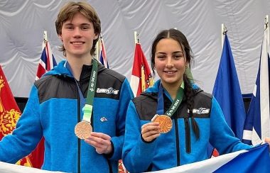 Dorian Thibault et Denise Thibault tiennent ensemble avec une chacun main le drapeau de la Nouvelle-Écosse. Dans leurs autres mains, ils tiennent les médailles bronzes autour de leurs cous. Ils sont dans leurs jackets de l'équipe de la Nouvelle-Écosse. En arrière-plan, on voit les drapeaux des provinces et térritoires du Canada.