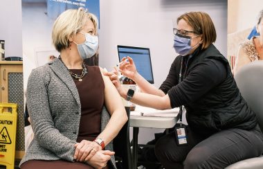 A woman wearing black gives a vaccine shot to Dr. Bonnie Henry in her arm in a medical clinic